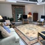 Cozy living room with a large ornate beige rug, brick wall, and a teal vintage mirror between chairs and a cabinet.